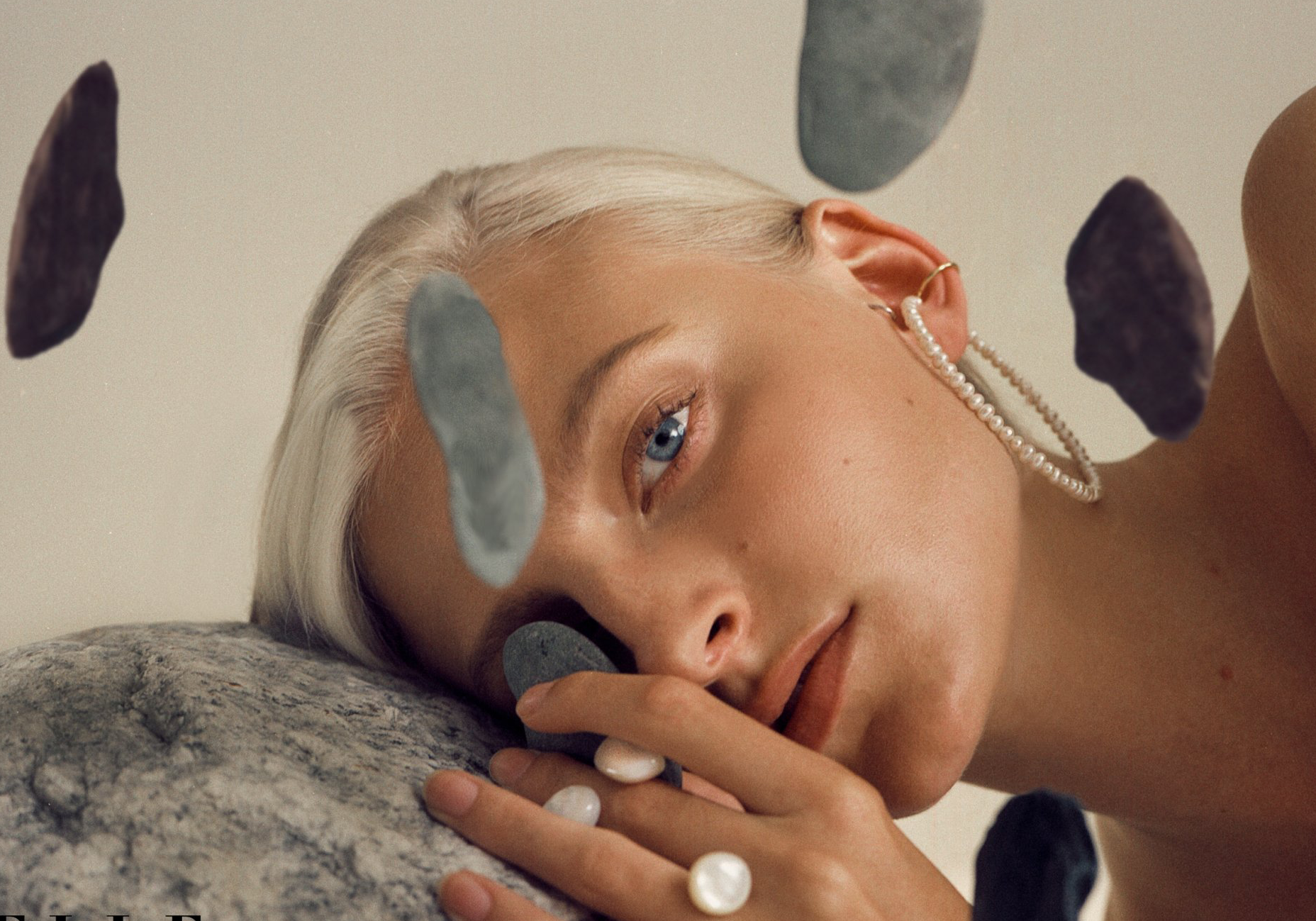 Blonde Frau mit blauen Augen ruht auf einem Stein, umgeben von schwimmenden Felsen. Sie trägt Creolen und einen Ring.