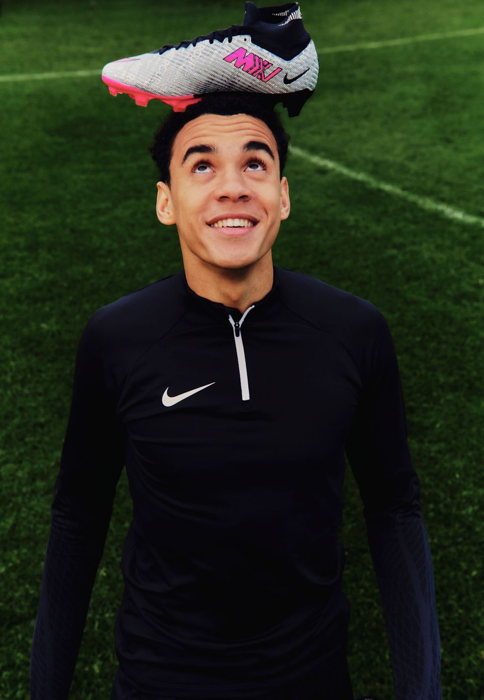 Mann balanciert einen Fußballschuh auf seinem Kopf und lächelt auf einem grünen Feld.