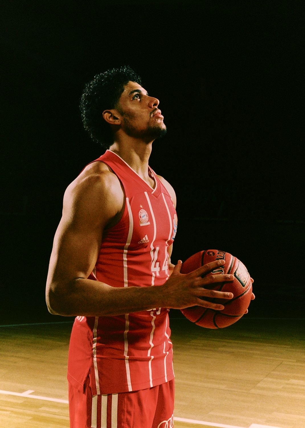 Basketballspieler im roten Trikot hält Ball, blickt nach oben, Spielfeld im Hintergrund.
