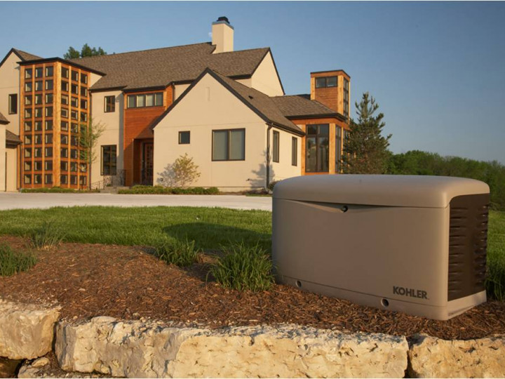 A beige generator sits in front of a modern house on a grassy lawn.