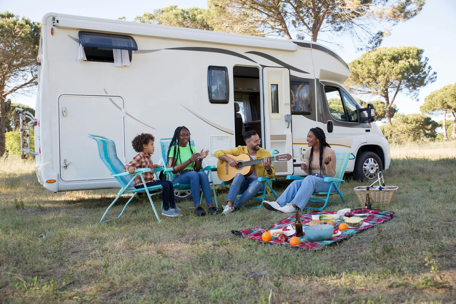 Family camping next to RV: singing, playing guitar, picnic. Blue chairs, blanket, food, and trees.