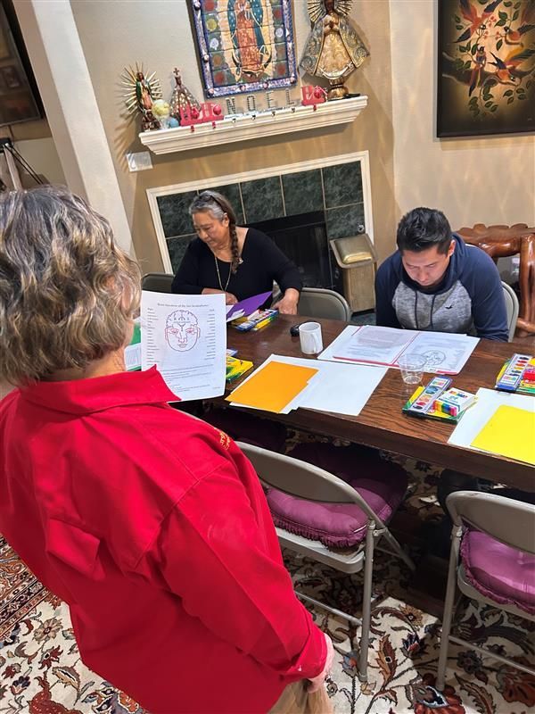 Three people sit at a table in a home, working on drawings on papers, while one stands holding a page.