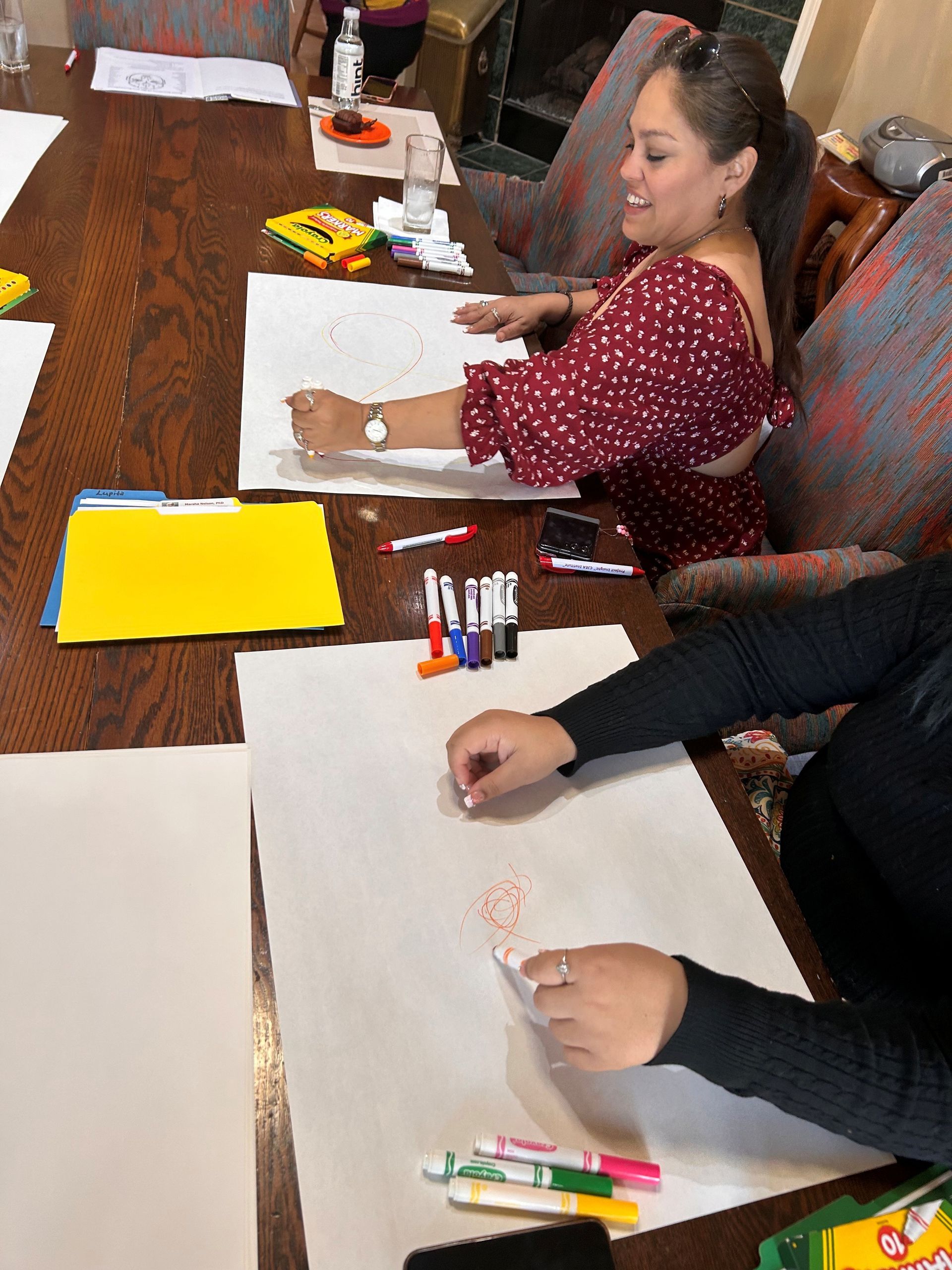 Two people sitting at a wooden table, drawing on large sheets of white paper with markers and crayons.