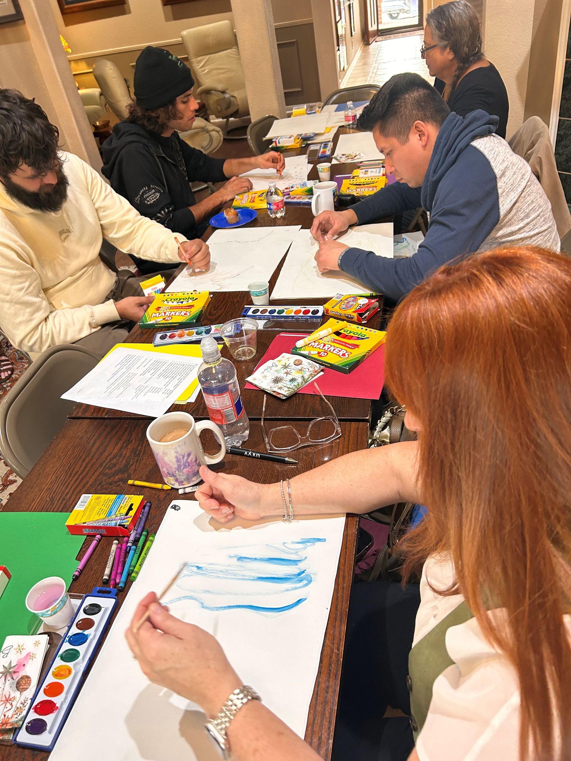 A group of people sit at a table, drawing and painting with watercolors and crayons in a workshop setting.