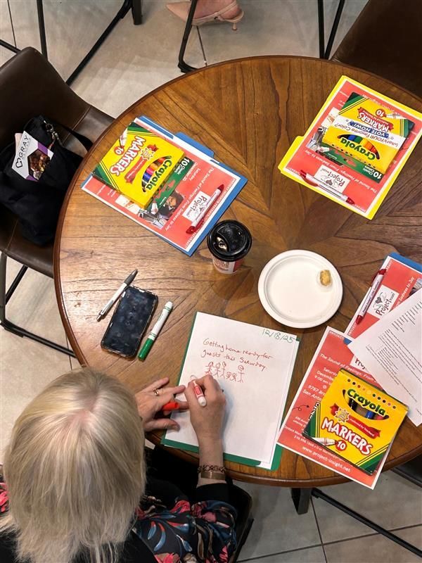 A person writing on a paper at a round wooden table with Crayola art supplies and a small white plate.