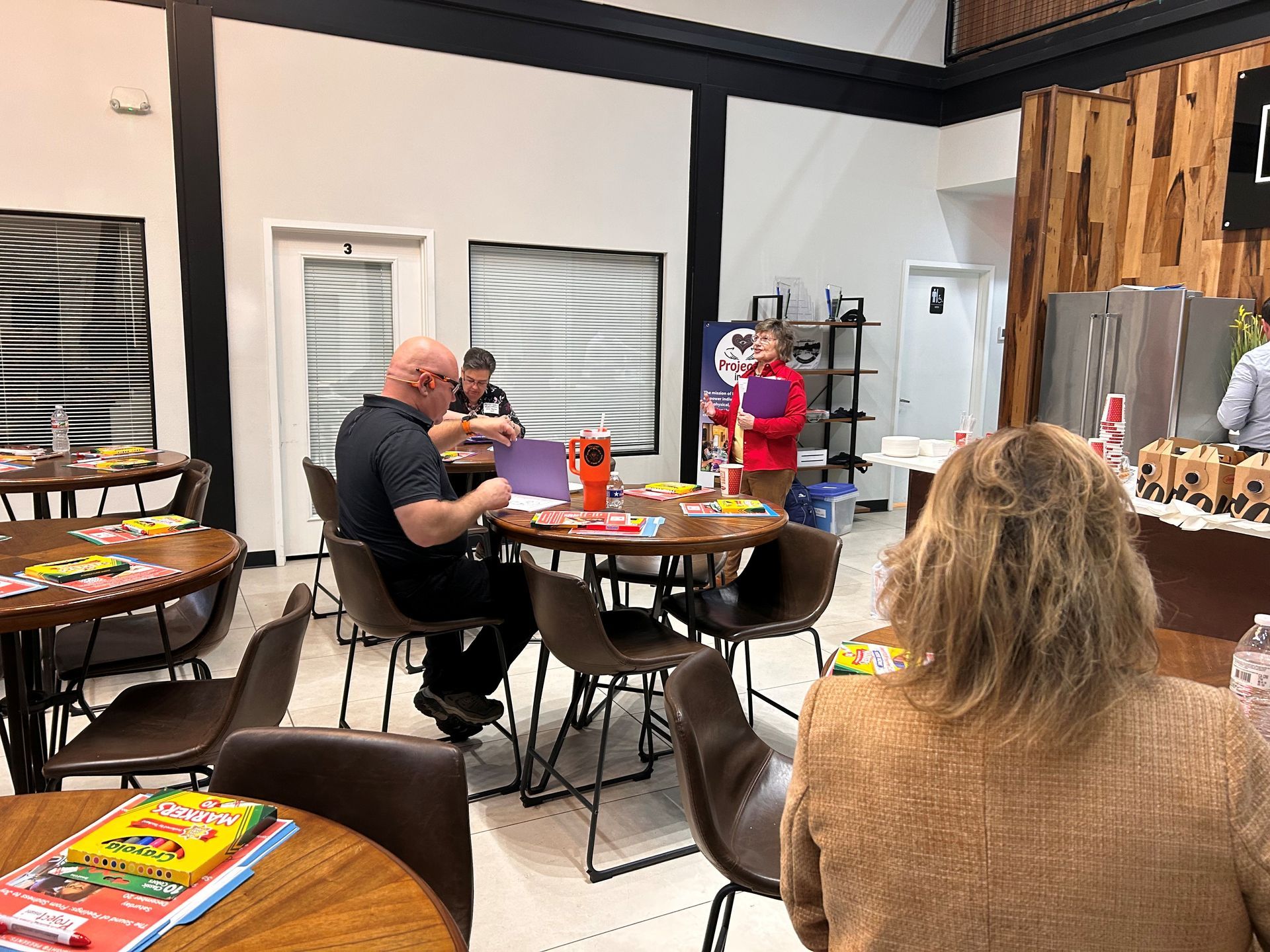 People sit at round tables in a bright, modern room, working on projects and talking during an informal group meeting.