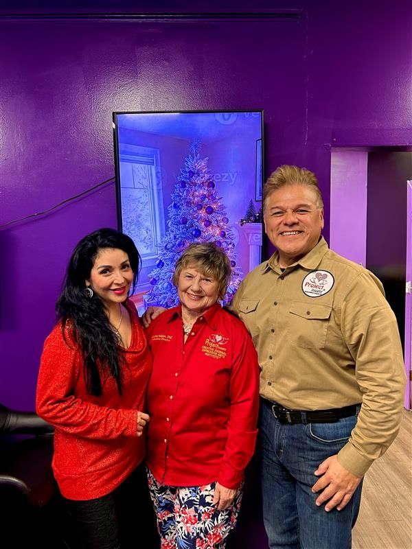 Three people smile while standing in front of a monitor showing a decorated Christmas tree against a purple wall.