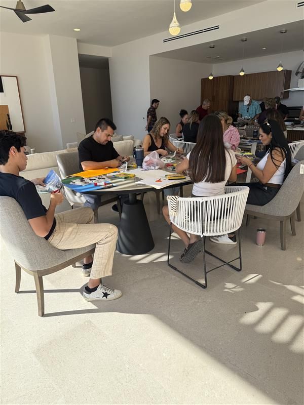 A group of people are seated at a circular dining table in a bright, open-plan room, working on art projects.