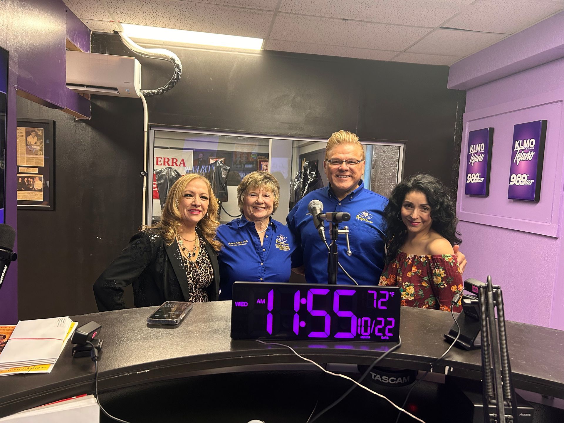 Four people pose for a photo behind a studio desk with a digital clock displaying 11:55 in a room with purple accents.