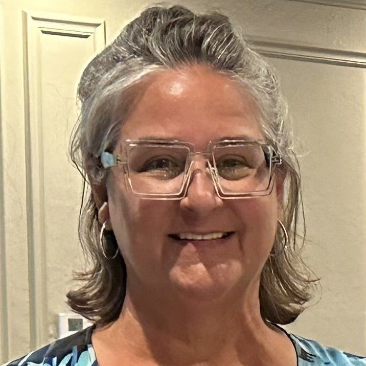 A smiling person with shoulder-length gray hair wears clear-framed glasses and silver hoop earrings.