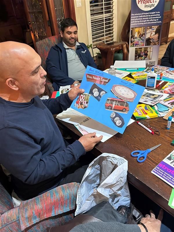 Two people sit at a table, one holding a blue poster board with images of a red bus and other objects for a group project.