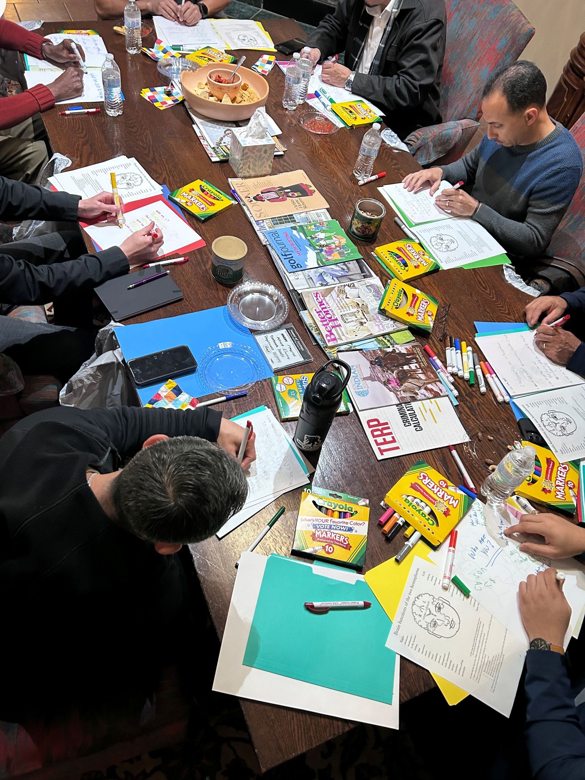 People sit around a wooden table, drawing and coloring in booklets with crayons and markers.