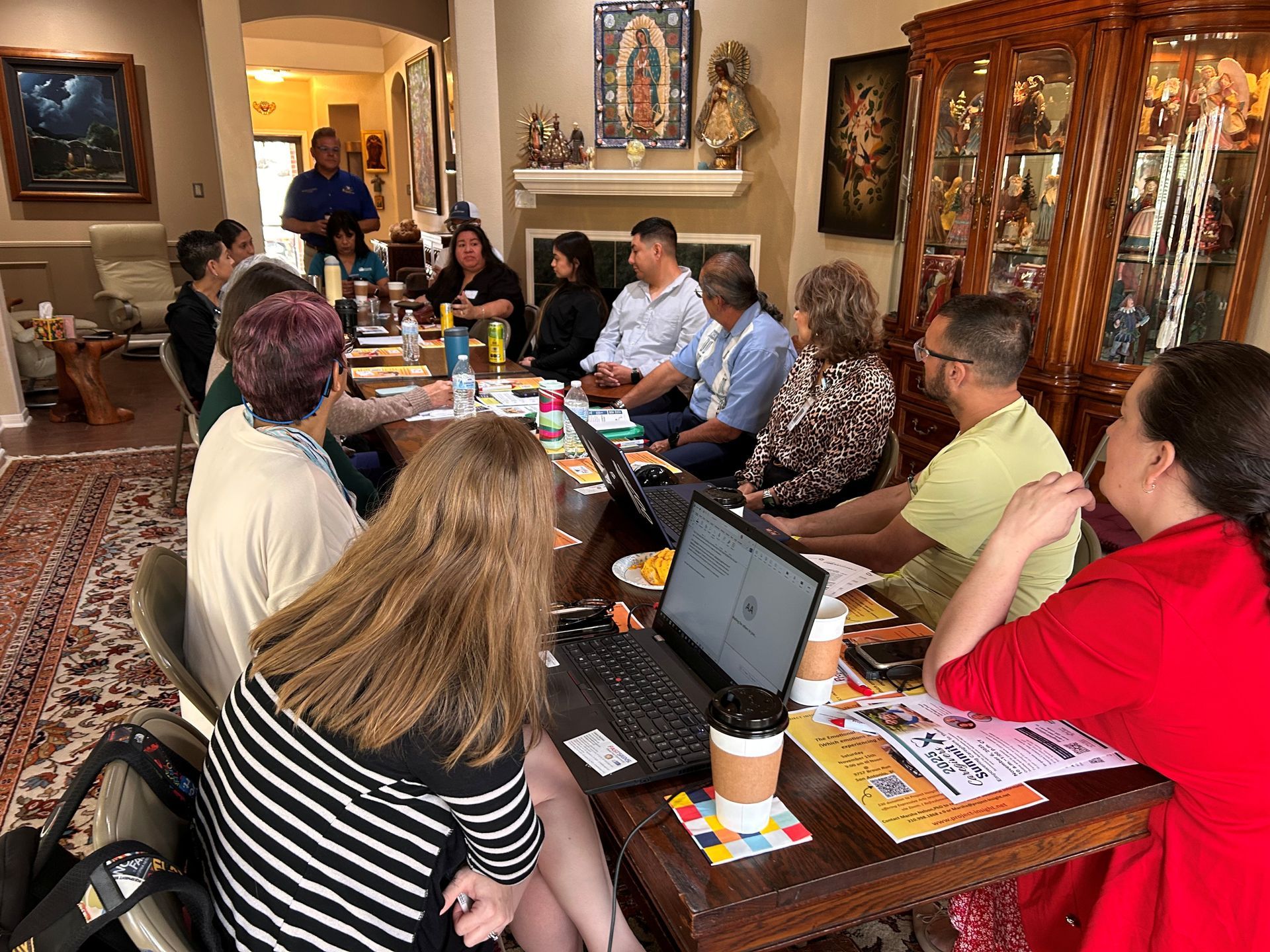 A group of people sitting around a table in a home setting for a meeting, with laptops and papers spread out.