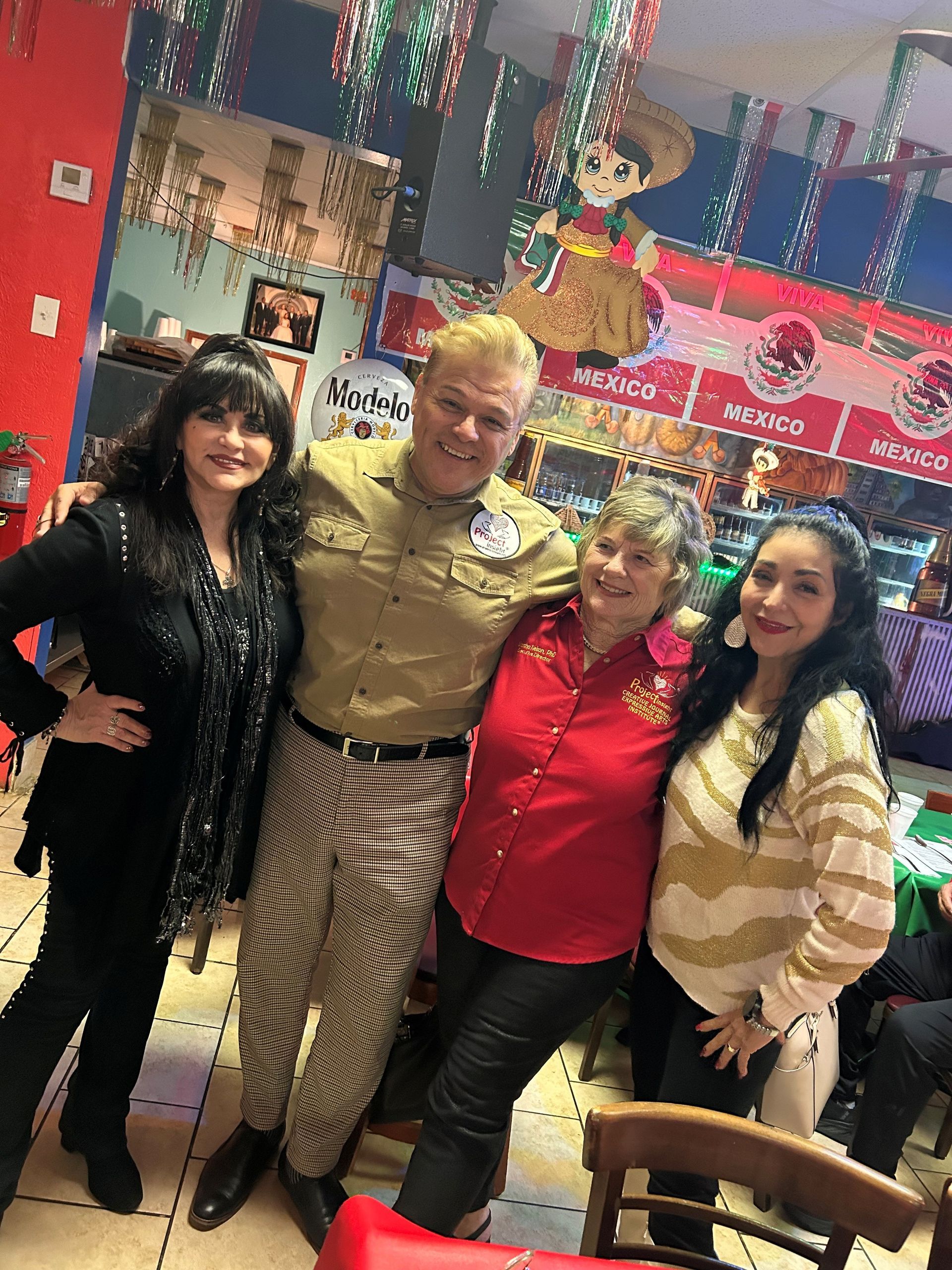 Four people pose together in a restaurant with festive decor and Mexican-themed wall art in the background.