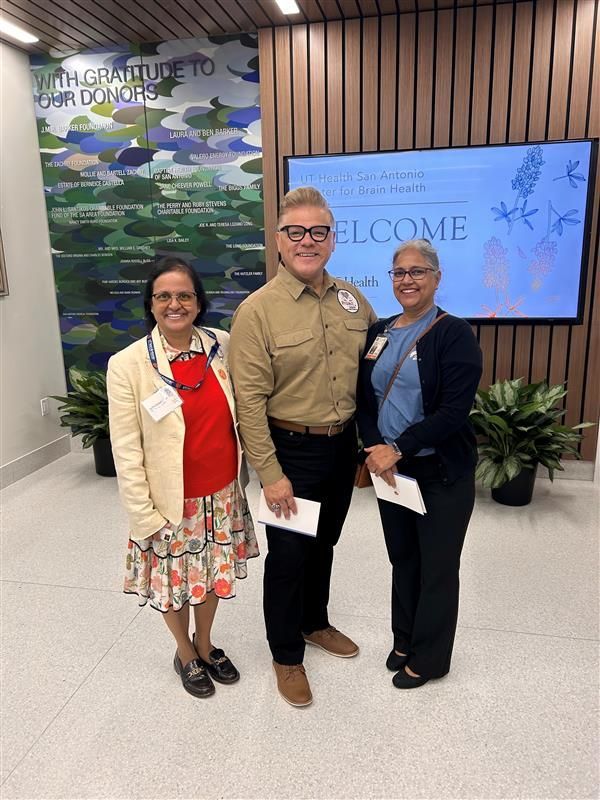 Three people stand smiling in front of a digital welcome sign and a mural labeled 