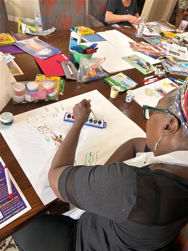 An individual uses a small watercolor set to paint on a large sheet of paper at a craft-filled wooden table.