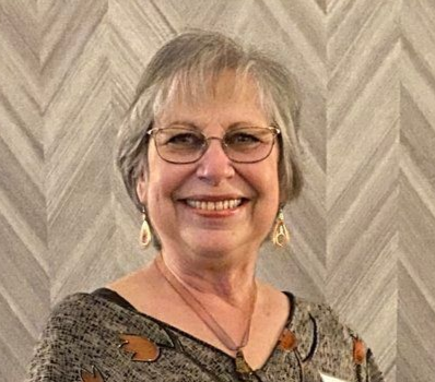Woman with glasses smiles in front of a patterned wall; wearing earrings, necklace, and floral shirt.