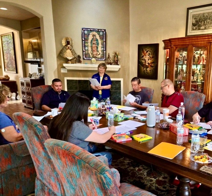 A group of people sit around a large dining table during a meeting in a home, with one person standing at the head.
