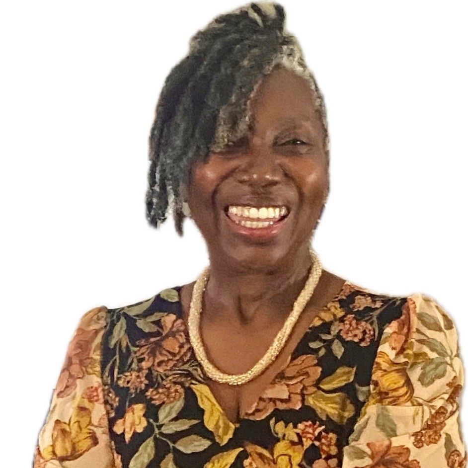 A person with dark locs smiles broadly, wearing a floral V-neck top and a beaded necklace against a white background.