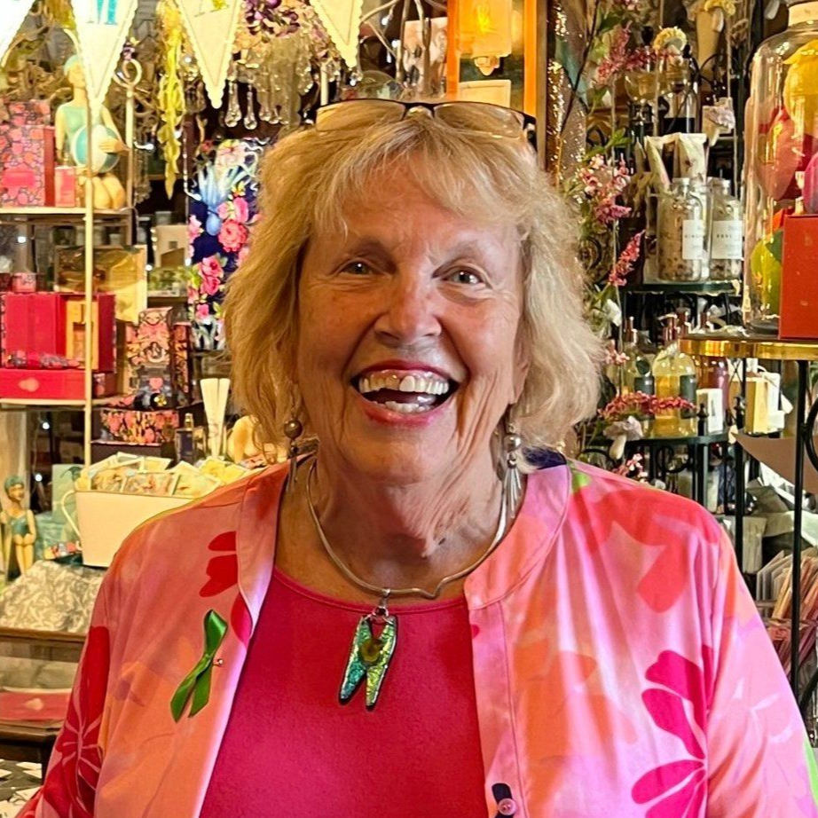 A smiling person with blonde hair wears a pink floral cardigan, bright pink shirt, and a green pendant in a gift shop.