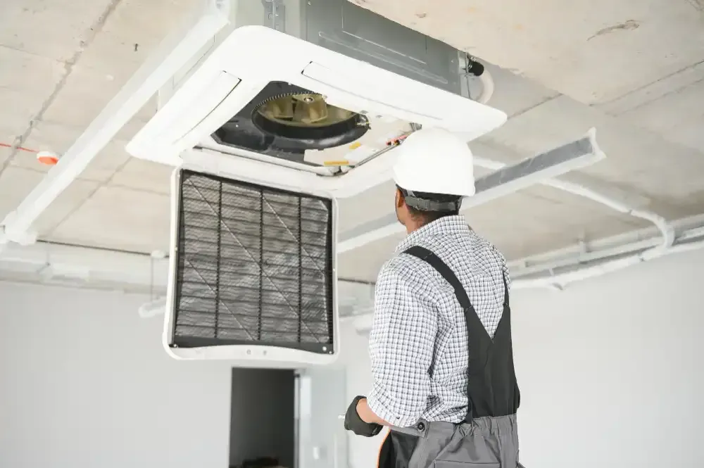 HVAC technician in white hard hat inspecting air filter of ceiling-mounted air conditioning unit.