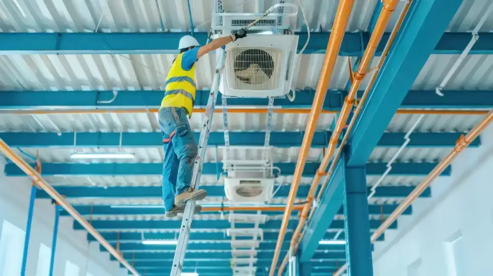 HVAC technician on a ladder, working on ceiling-mounted air conditioning unit in a factory.