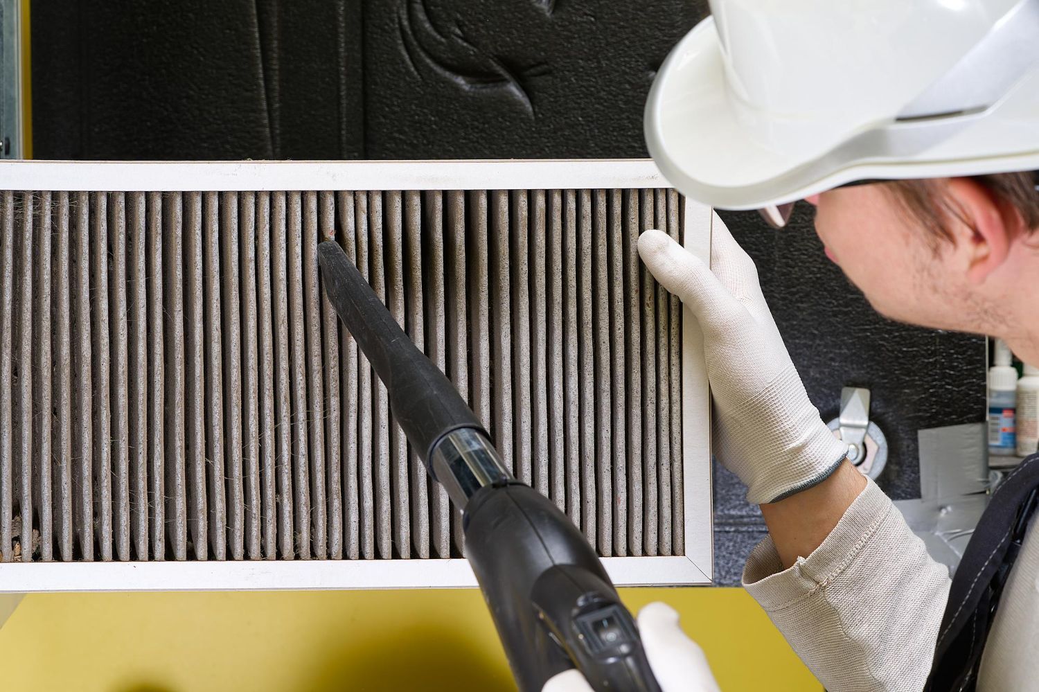 Person vacuuming a dirty air filter, wearing gloves and a hard hat.