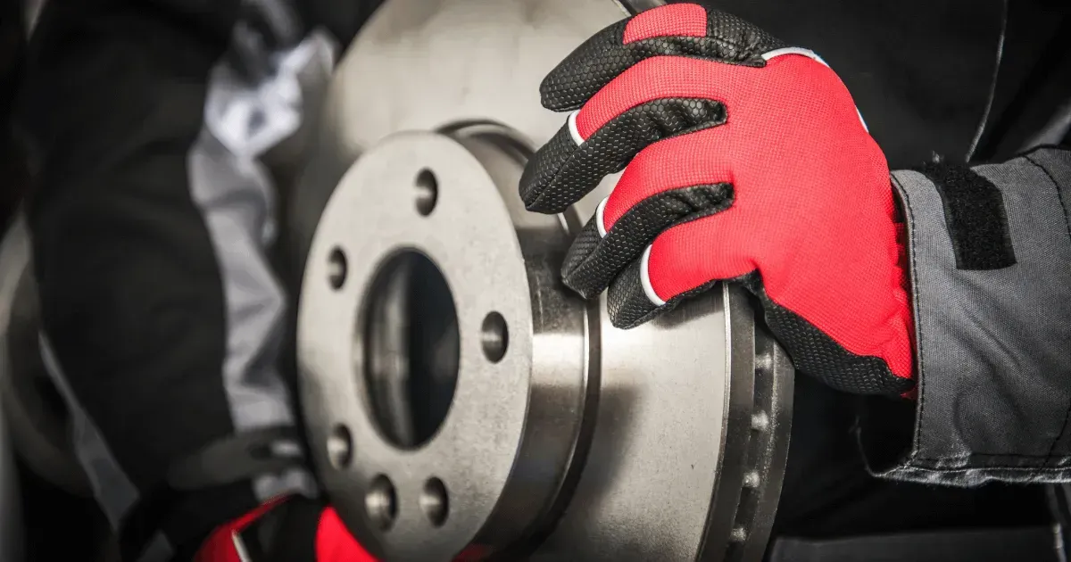 A mechanic wearing red and black work gloves holds a circular metal brake rotor.