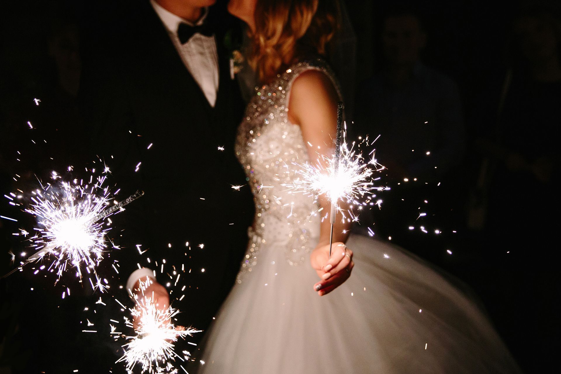 Couple kissing, holding sparklers at a nighttime wedding celebration, bride in white dress, groom in black suit.