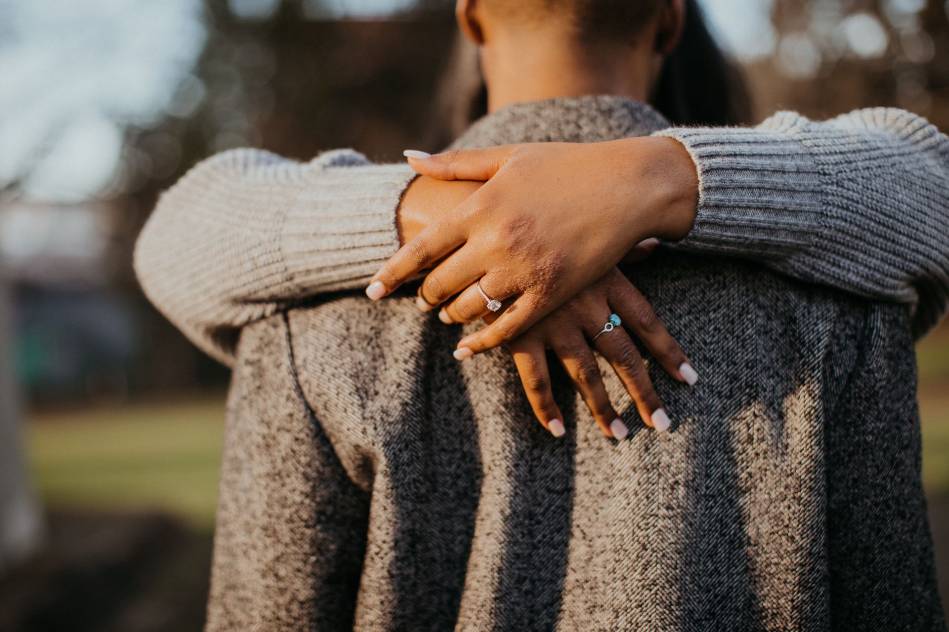 Arms crossed around a person's back; hands with rings visible. The person wears a gray coat in an outdoor setting.