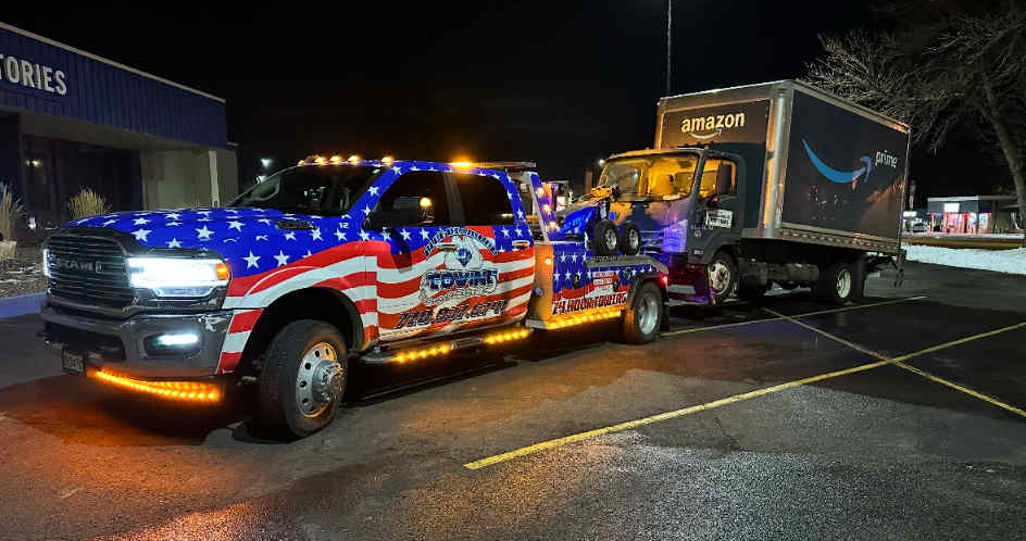 American flag-themed tow truck towing an Amazon delivery truck at night.
