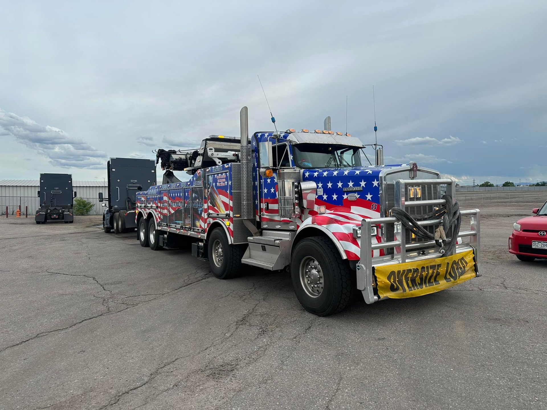 Patriotic tow truck hauling a vehicle, parked on asphalt, cloudy sky.