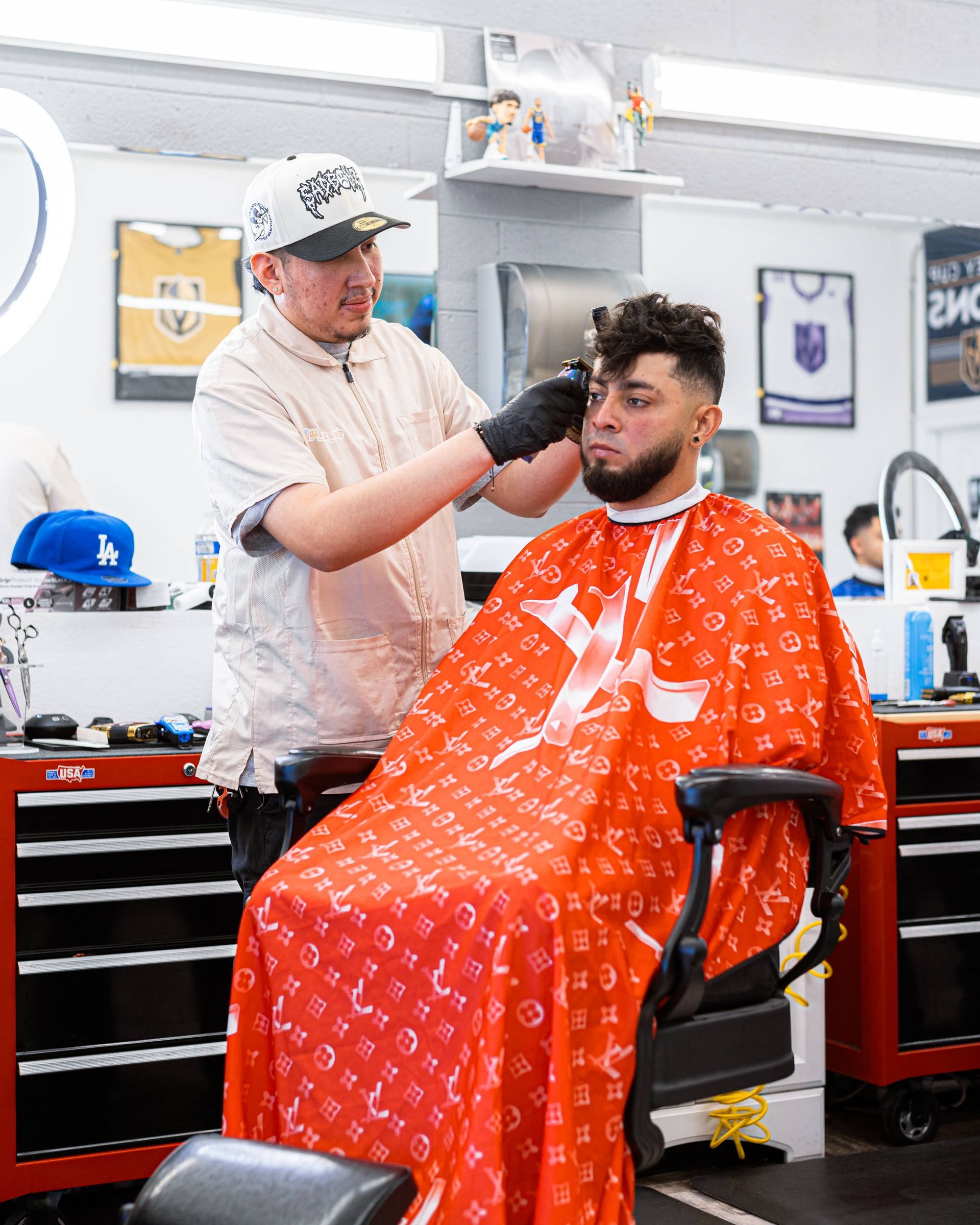 Barber trimming a seated client’s hair in a shop, with a red cape and black gloves.