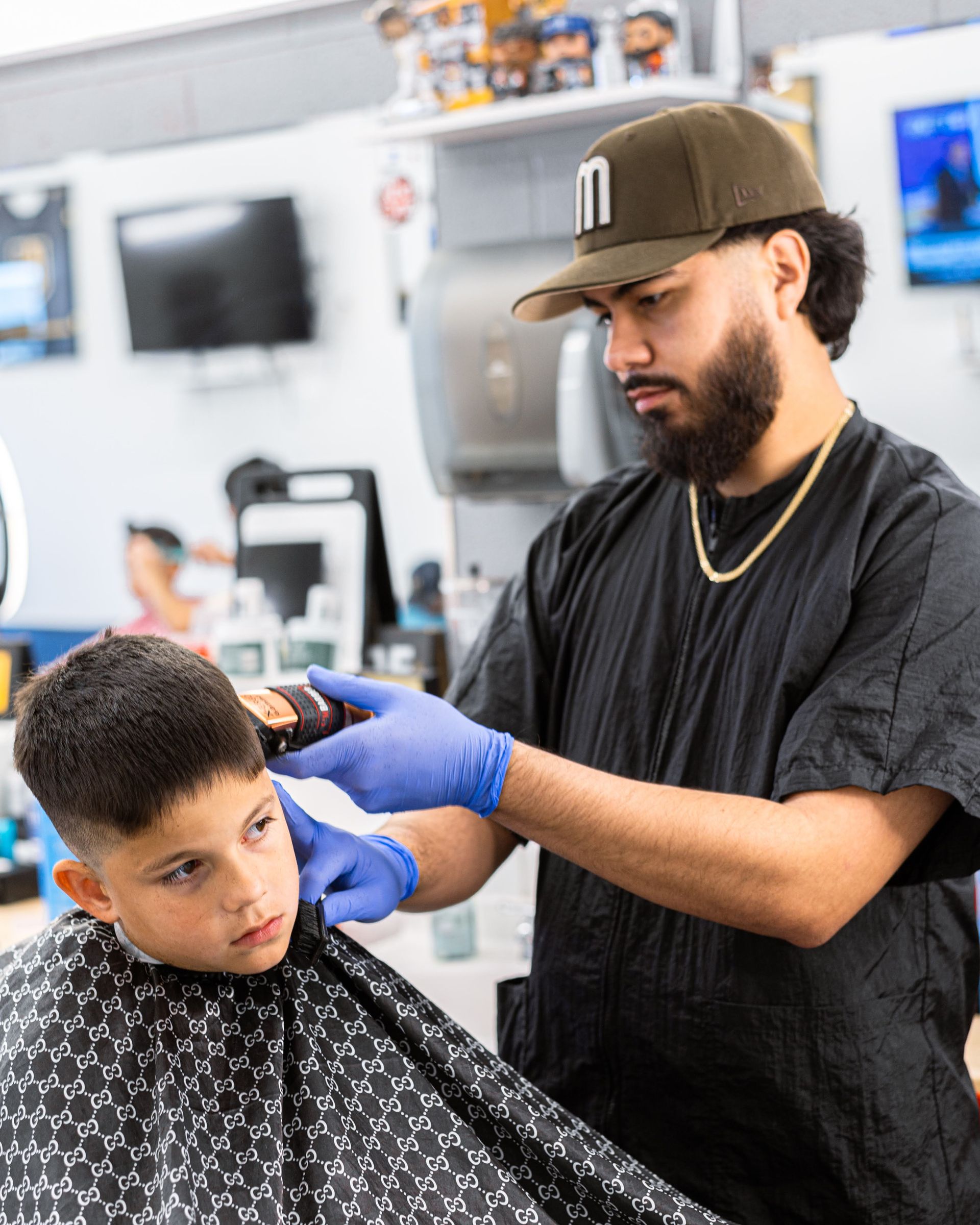 Barber trimming a client’s hair in a bright barbershop, wearing gloves and a cap.
