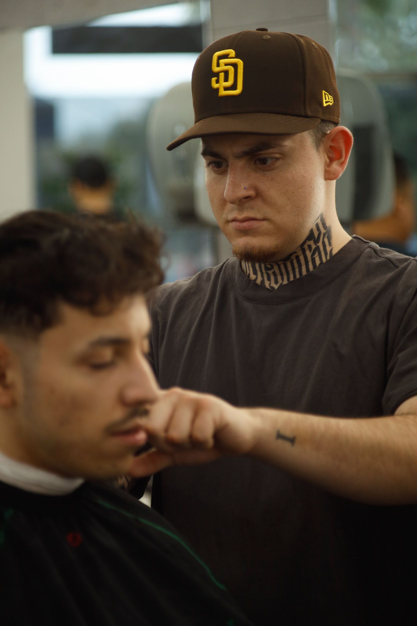 Barber trimming a customer’s beard in a shop, with a focused expression and a brown cap.