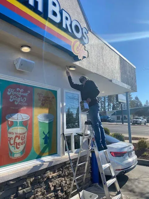 Man painting a Dutch Bros Coffee sign while standing on a ladder. Sunny day.