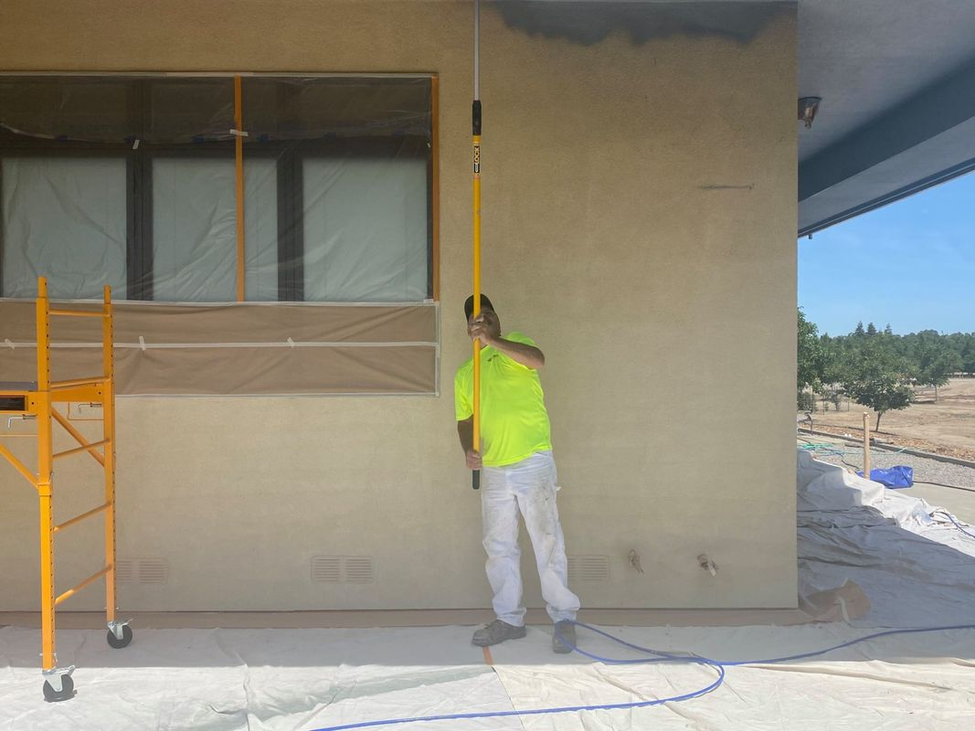 A person in a neon shirt painting an exterior wall with a long-handled roller. Yellow scaffolding stands nearby.