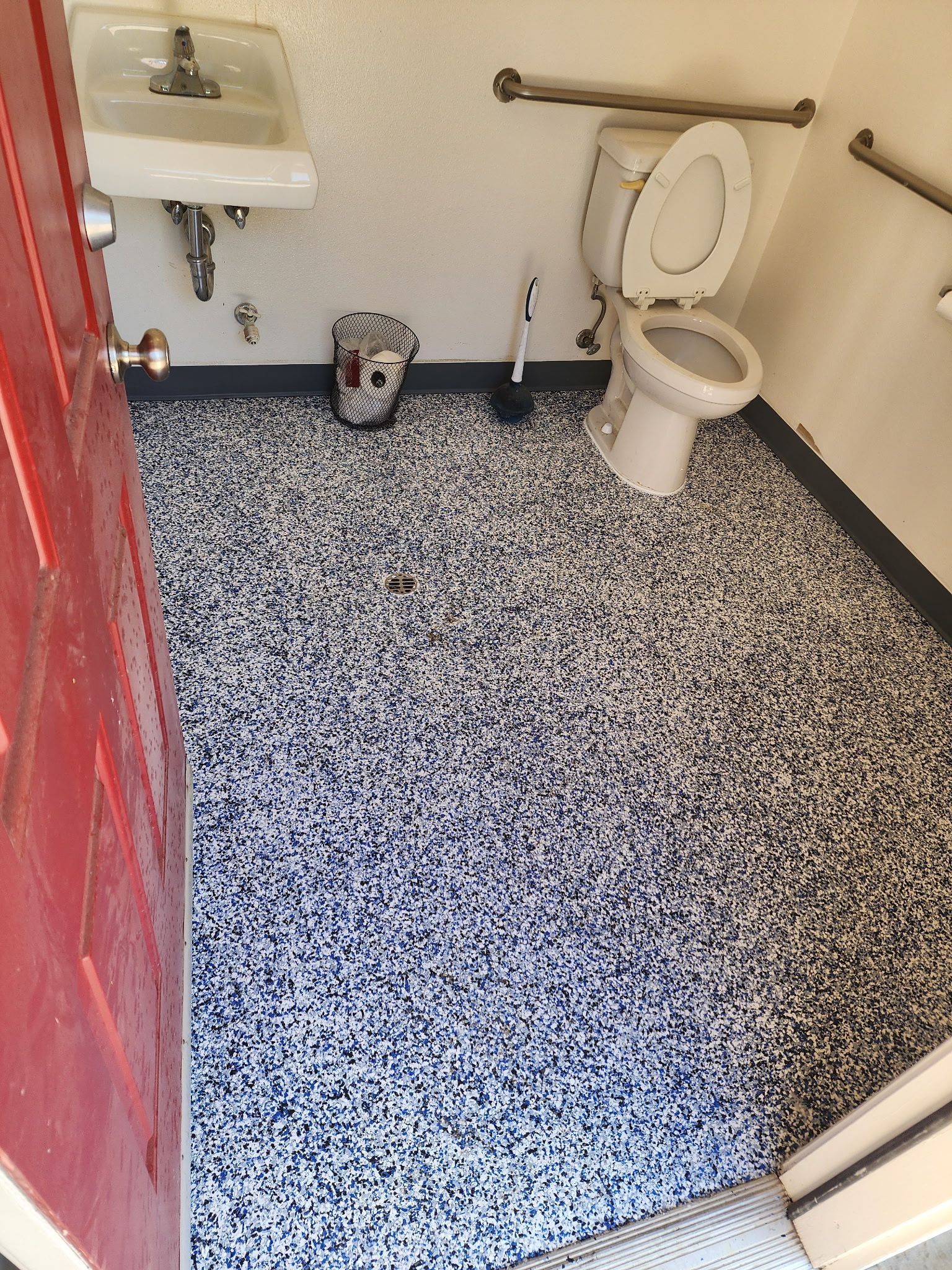 A restroom with a sink, a toilet, a grab bar, and a patterned black and white floor, viewed through an open red door.