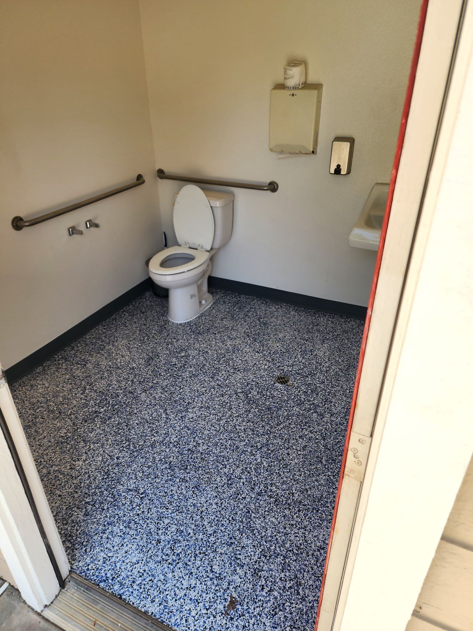 Accessible restroom stall with white toilet, handrails, toilet paper dispenser, and a speckled black and white floor.