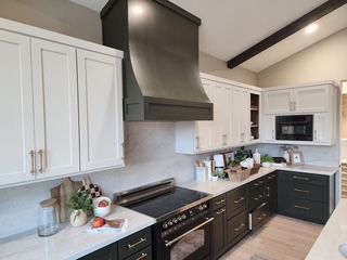 A modern kitchen featuring white upper cabinets, dark base cabinets, a custom vent hood, and a black range.