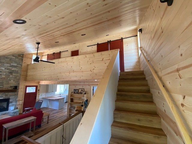Interior of a cabin with wooden walls, stairs, and loft. Red doors and a stone fireplace are visible.