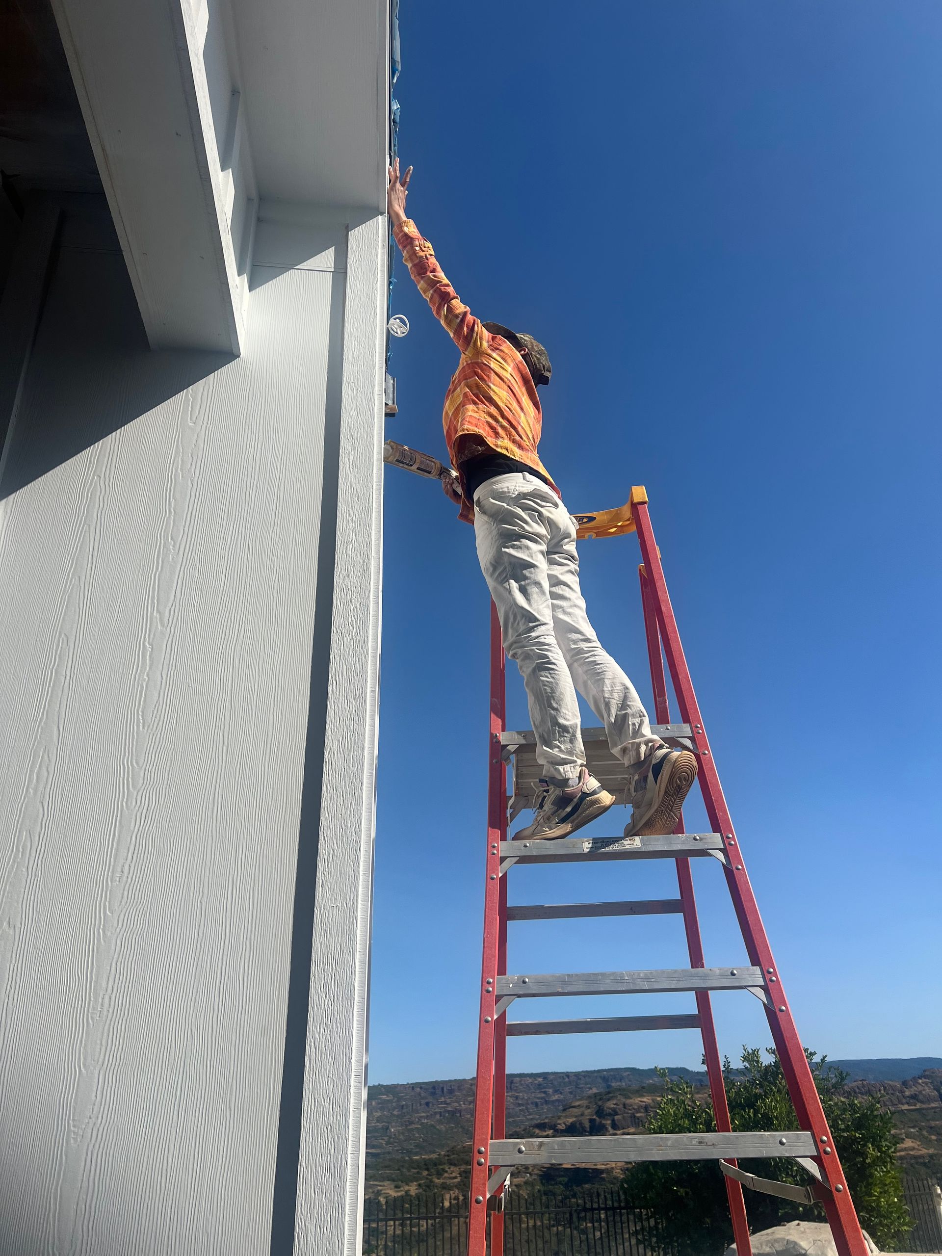A person wearing an orange shirt stands near the top of a red ladder, reaching up to touch the edge of a house roof.