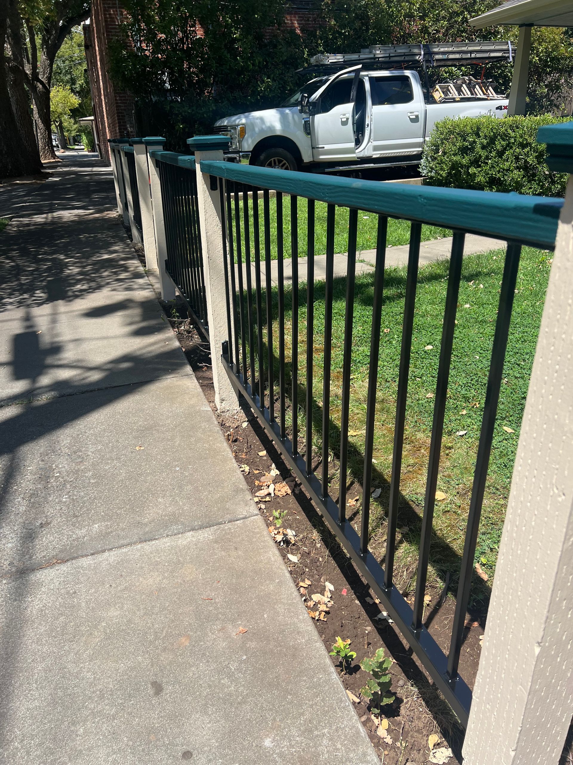 A black metal fence with a green top rail runs along a concrete sidewalk, with a white truck parked in the background.