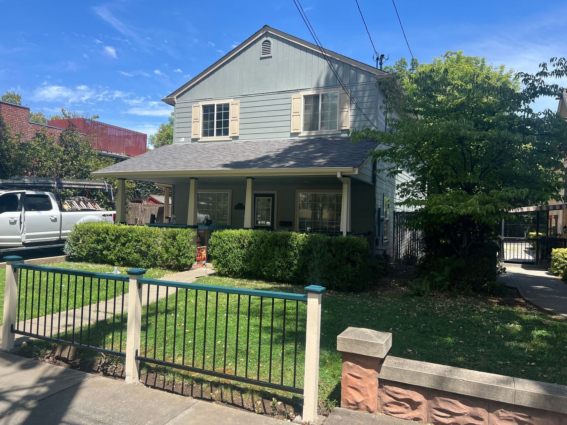 A two-story grey house with a porch and green hedge sits behind a black and green metal fence on a sunny day.