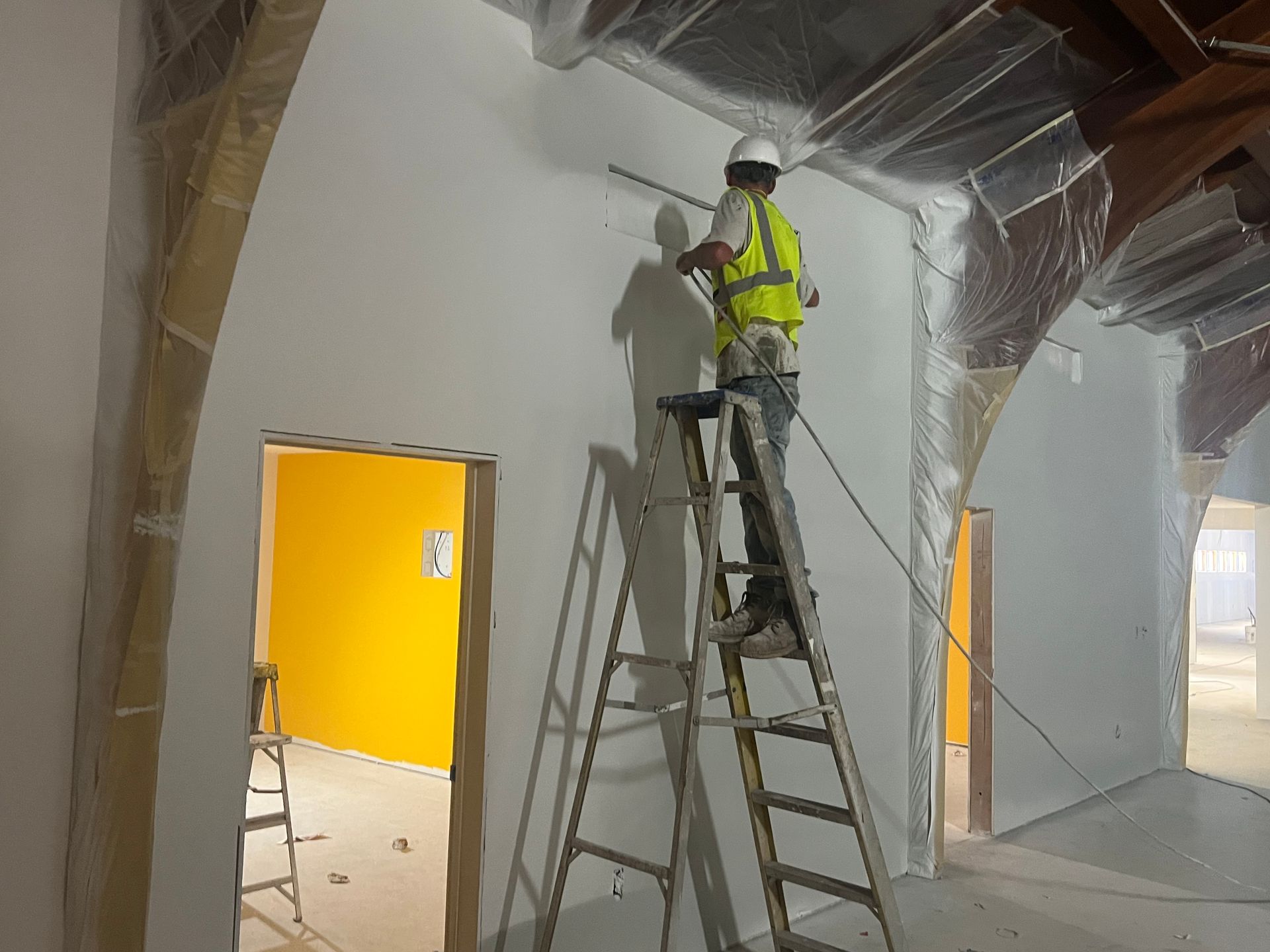 A construction worker wearing a hard hat and high-visibility vest stands on a stepladder while working on a white wall.
