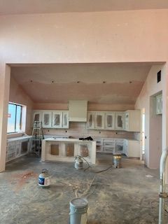 A wide-angle view of a kitchen under construction with white cabinets, a central island, and freshly plastered walls.