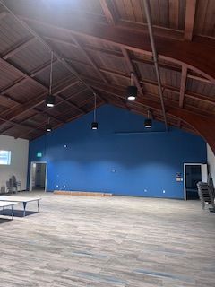 A large, empty hall with a blue wall, exposed wooden beamed ceiling, pendant lights, and light-colored flooring.