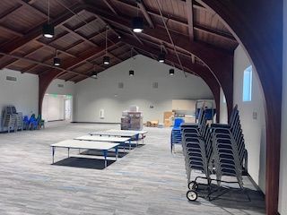 Spacious hall with high wood-beamed ceilings, grey carpet, arranged tables, and stacks of chairs on wheels.