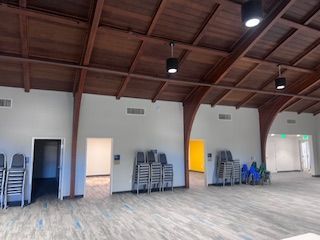 A large, empty hall with a high wood-beamed ceiling, grey carpet, white walls, multiple doorways, and stacks of chairs.