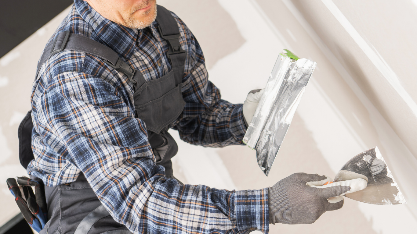 Man in plaid shirt and work gloves uses a trowel to apply drywall mud to a wall.